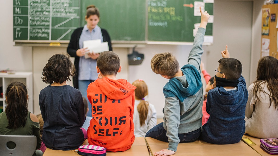 Schüler melden sich während eine Lehrerin in einem Klassenzimmer vor einer Tafel steht.