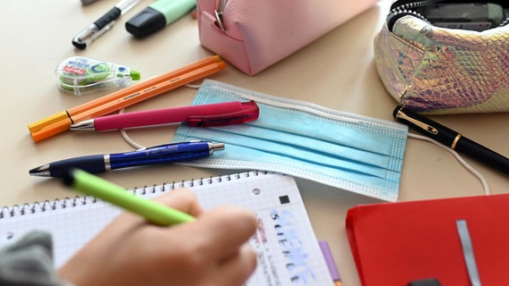 Auf einem Schreibtisch liegen Stifte und ein Mund-Nasen-Schutz. Eine Kinderhand schreibt. © picture alliance/dpa Foto: Uli Deck