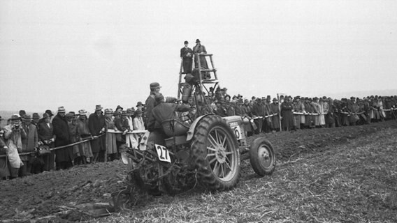 Ein schwarz-weißes Foto von zwei Männern auf einem Traktor, die vor Zuschauern pflügen. © NDR Foto: Bernhard Glasow
