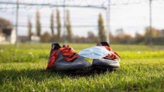 Zwei Fußballschuhe und ein Mund-Nasen-Schutz liegen auf einem Fußballrasen. © picture alliance / HMB Media Foto: Heiko Becker