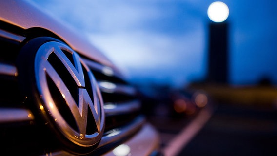 Das Logo eines VWs ist im Vordergrund zu sehen. Im Hintergrund leuchtet der VW-Turm unscharf. © dpa - Bildfunk Foto: Julian Stratenschulte
