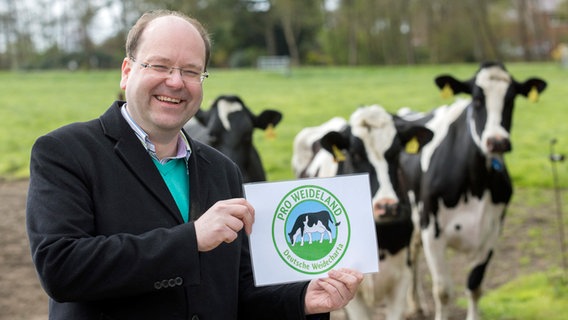 Landwirtschaftsminister Christian Meyer (Grüne) steht auf einer Kuhweide und hält das neue "Pro Weideland"-Siegel in der Hand. © dpa Bildfunk Foto: Jörg Sarbach