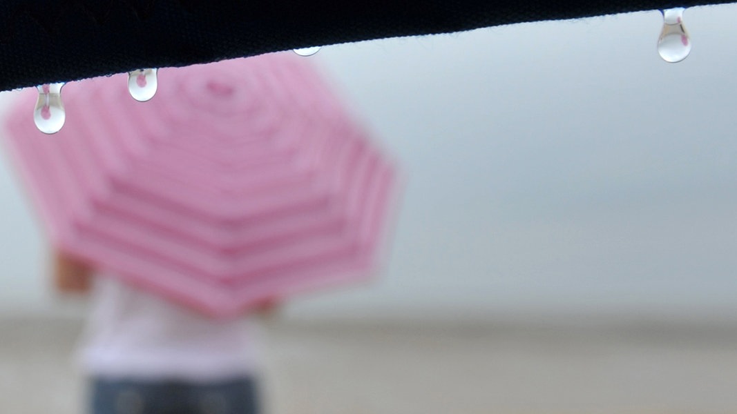 Eine Frau steht mit einem Regenschirm geschützt vor einem Strandkorb am Strand.