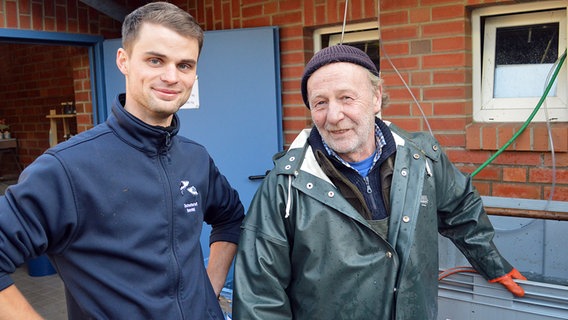 Der junge Teichpächter (l.) steht neben einem älteren Mann (r.) vor einem Gebäude in Reinfeld. © NDR Foto: Anne Passow