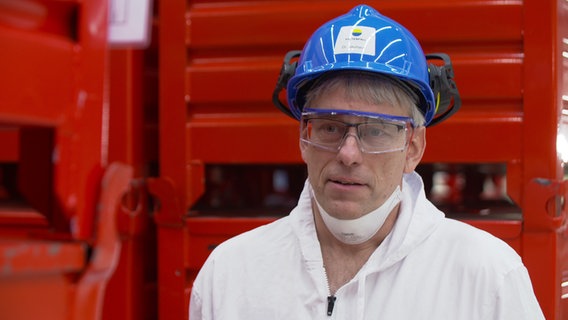 Ein Mann mit einem blauen Helm und einer Schutzbrille steht vor einem roten Container und blickt in die Kamera. © NDR 