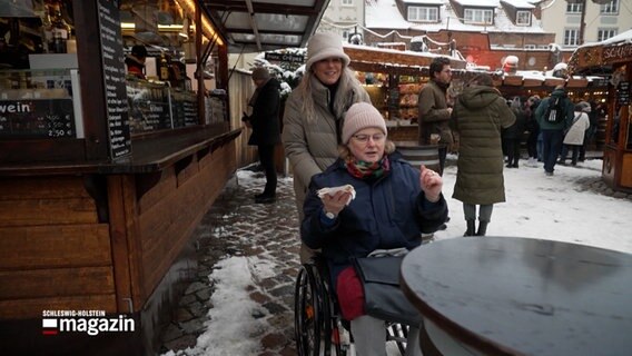 Eine Rollstuhlfahrerin wird über den Weihnachtsmarkt geschoben. © NDR 