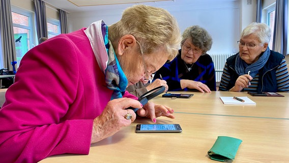 Eine Frau betrachtet das Display eines Smartphones, das vor ihr auf dem Tisch liegt, mit einer Lupe. © NDR Foto: Fabian Boerger