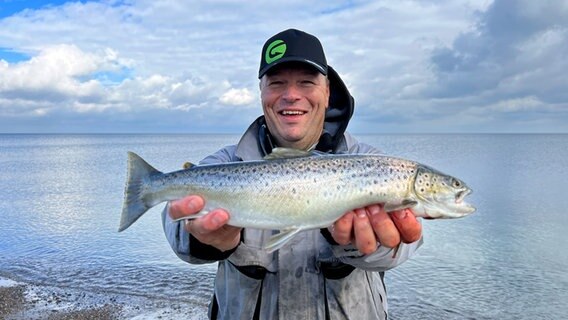 Danny Hrubesch steht vor der Ostsee und hält eine gefangene Meerforelle in den Händen. © NDR Foto: Lena Storm