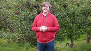Nicho Nölting steht vor einem Apfelbaum und hält einen aufgeschnittenen Apfel in der Hand.  Foto: Andrea Ring