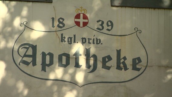 Ein Schild mit der Überschrift "1839 kgl. priv. Apotheke" hängt an einem Gebäude. © NDR 