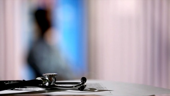 Ein Stethoskop liegt auf einem Tisch im Vordergrund, im Hintergrund sind die verschwommenen Umrisse einer Person zu erkennen. © NDR 