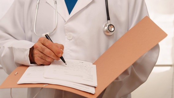 Arzt in weißem Kittel und mit Stethoskop schreibt etwa auf ein Blatt in einem Ordner. © imageBROKER/josexhernandez Foto: jose hernandez