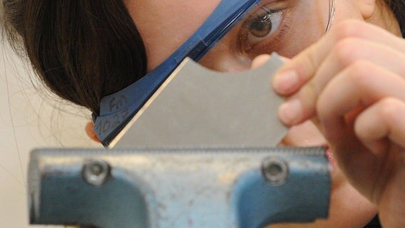 Eine Auszubildende trägt eine blaue Schutzbrille und steckt ein Stück Metall in eine Vorrichtung. © picture alliance / dpa Foto: Marcus Brandt