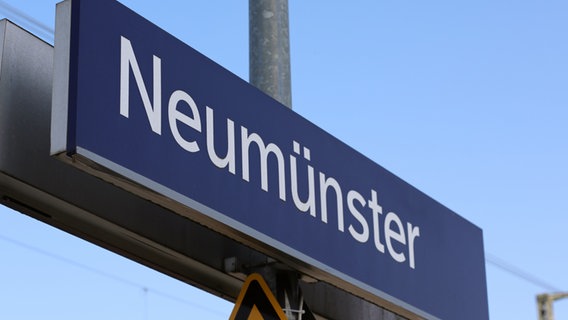 Das blaue Schild mit der Überschrift "Neumünster" hängt an einem Mast an einem Bahnsteig am Bahnhof in Neumünster. © NDR Foto: Pavel Stoyan