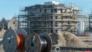 Schleswig, ein Rohbau mit Baugerüsten auf einer Baustelle. Im Vordergrund zwei große Kabeltrommeln aus Holz mit Erdkabeln für Glasfaser und Strom. © picture alliance / SULUPRESS.DE Foto: Torsten Sukrow