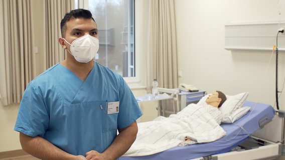 Der auszubildende Krankenpfleger Mohammad Berri steht mit einem Mundschutz vor einem Bett, in dem eine Puppe liegt. © NDR 