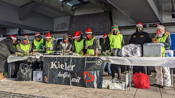 Menschen mit Weihnachtsmützen stehen hinter Tischen auf denen Essen, Trinken und Tannengrün liegt und steht. © NDR Foto: Balthasar Hümbs