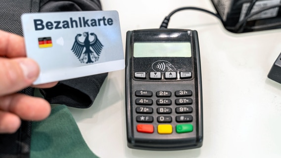 Jemand hält eine EC-Karte mit der Aufschrift "Bezahlkarte" vor ein Kartenlesegerät. © IMAGO / Bihlmayerfotografie 