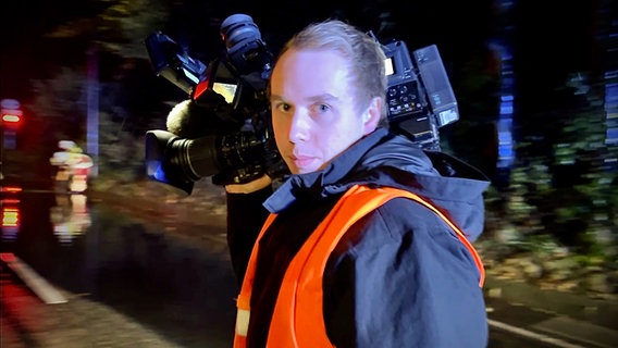 Ein Kameramann hält eine große Fernsehkamera auf der Schulter und blickt in die Kamera bei Nacht. © NDR Foto: Christoph Deuschle