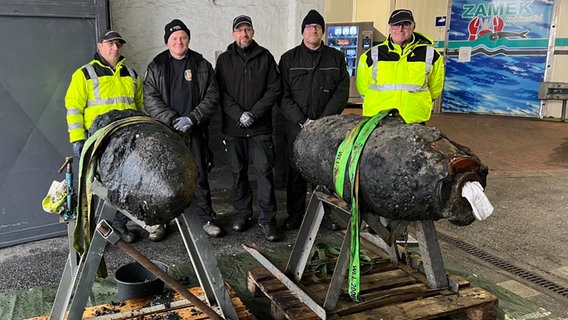 Fünf Mitarbeiter des Kampfmittelräumdienstes stehen hinter zwei entschärften Fliegerbomben in einer Halle auf dem Kieler Ostufer. © NDR Foto: Sofia Tchernomordik