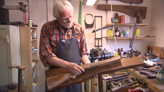Ein Büchsenmacher steht in einer Werkstatt und zeigt eine im Bau befindliche Waffe.  