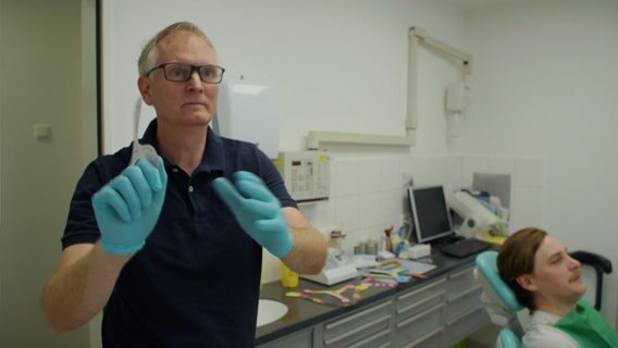 Torsten Gründel wirft in seiner Praxis einen Bumerang links im Bild. Während ein Patien im Behandlungsstuhl liegt recht im Bild. © NDR 