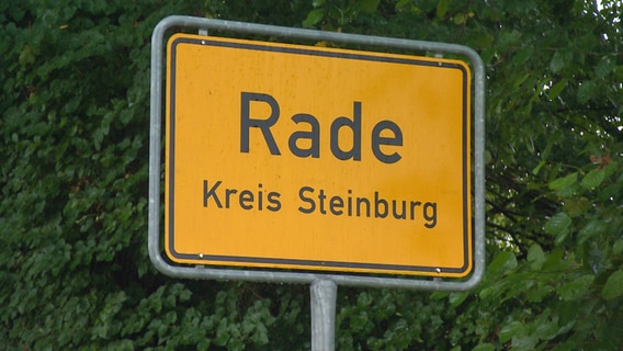 Ein gelber Ortsschild von Rade im Kreis Steinburg steht vor grünen Bäumen. © NDR 