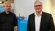 Sönke Rix (r.) steht neben dem Moderator Konstantin Gill (l.) in einem Sendestudio des NDR in Kiel neben einem NDR-1-Welle-Nord-Banner und lächelt dezent in die Kamera. © NDR Foto: Pavel Stoyan