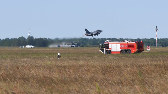 Ein Fahrzeug der Flughafenfeuerwehr steht auf dem Rollfeld. Im Hintergrund startet eine amerikanische F-16. © NDR Foto: Jörn Zahlmann