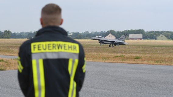 Ein Mann steht auf einem Rollfeld und betrachtet ein ankommendes Militär-Flugzeug. Auf seinem Rücken befindet sich der Schriftzug "Feuerwehr". © NDR Foto: Jörn Zahlmann