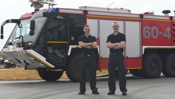 Zwei Feuerwehrleute posieren vor einem Feuerwehr-Löschfahrzeug der Bundeswehr. © NDR Foto: Jörn Zahlmann