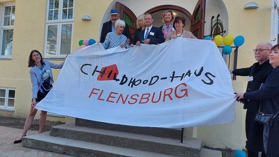 Mehrere Personen, darunter Fraktionsvorsitzende der Grünen Eka von Kalben, halten einen Banner vor dem Eingang in eine Villa in Flensburg bei der Eröffnung des Childhood-Houses. © NDR Foto: Laura Albus