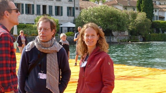 Zwei Männer und eine Frau aus dem Team, die die "Floating Piers" von Christo mit Stoff eingehüllt haben, stehen auf der Kunstinstallation und machen Pause. © NDR Foto: Julian Marxen und Hauke von Hallern