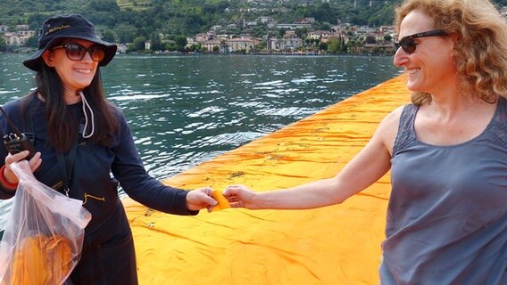 Eine Helferin gibt einer Besucherin der "Floating Piers" von Christo ein Stück des Stoffes, mit dem die Piers eingehüllt sind. © NDR Foto: Julian Marxen und Hauke von Hallern