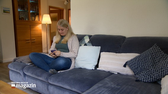 Eine junge Frau sitzt auf der Couch und blickt auf ein Handy. © NDR 