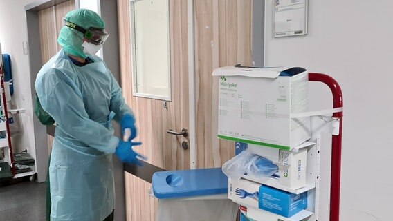 Eine Pflegekraft in Vollmontur zieht blaue Gummihandschuhe vor einem Patientenzimmer an. © NDR Foto: Cassandra Jane Arden