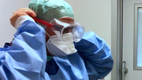 Krankenpfleger Dirk Zöllner (l.) aus Itzehoe und ein Kollege stehen im blauen Kittel und mit Mundschutz vor der Tür eines Patientenzimmers und setzen Schutzbrillen auf. © NDR Foto: Screenshot