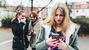 Ein Mädchen steht im Hof einer Schule und schaut auf ihr Smartphone, während im Hintergrund zwei andere Mädchen lachen. © picture alliance / klicksafe / Maribelle Photography Foto: klicksafe/Maribelle Photography