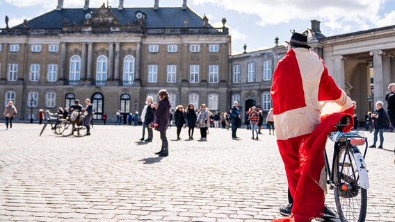Ein Mann, umhüllt von einer dänischen Flagge, schiebt sein Fahrrad über einen belebten Platz vor einem Schloss. © picture alliance / Ritzau Scanpix Foto: Niels Christian Vilmann