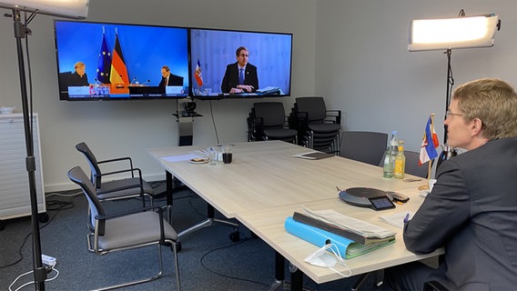 Daniel Günther sitzt einem großen Tisch und guckt auf zwei Bildschirme, auf dem die Pressekonferenz sowie er zu sehen sind. © NDR Foto: Julia Stein