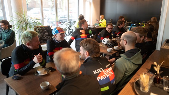 Fahrradfahrerinnen und -fahrer sitzen in einem Café zusammen. © NDR Foto: Lisa Pandelaki