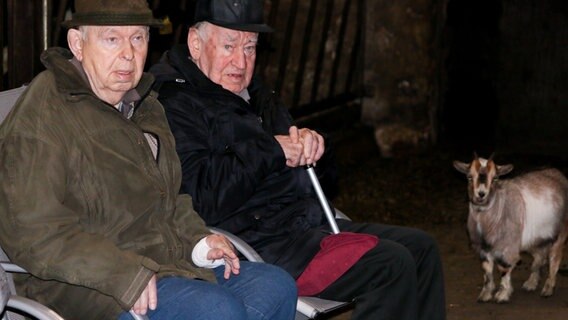 Zwei ältere Männer mit Hut sitzen in einem Stall,  neben ihnen steht eine kleine Ziege.  Foto: Claudio Campagna