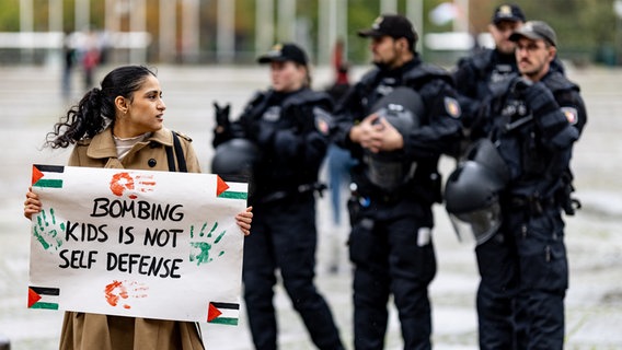 Eine Frau mit einem Plakat mit der Aufschrift "bombing kids is not self defence" steht vor Polizeikräften auf dem Rathausmarkt in Kiel, nachdem eine propalästinensische Demo in Kiel verboten wurde. © dpa-Bildfunk Foto: Axel Heimken