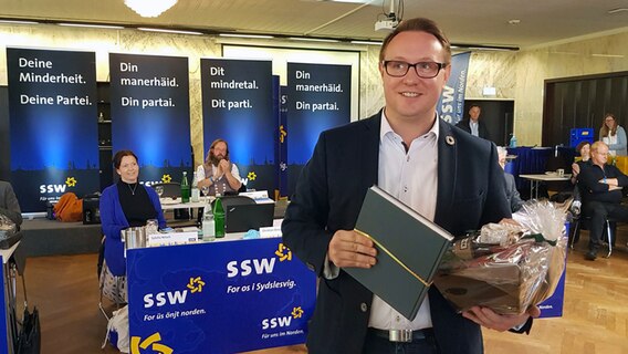 SSW-Politiker Christian Dirschauer steht mit einem Buch und eine Korb in den Händen nach der Wahl zum Parteichef mitten in einem Raum und lächelt. © NDR Foto: Lukas Knauer