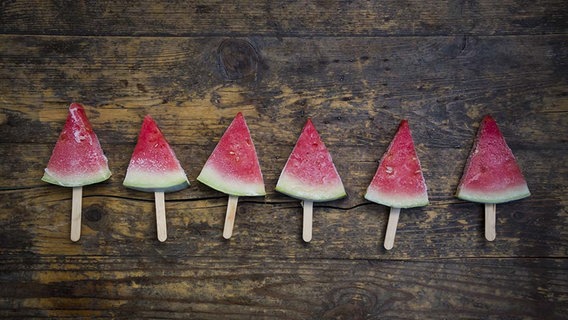 Dreieckige Stücke einer Melone wurden auf einen Stiel aufgespießt und eingefroren. Mehrere davon liegen in Reihe auf einer schwarzen Schieferplatte. © imago/Westend61 Foto: imago/Westend61