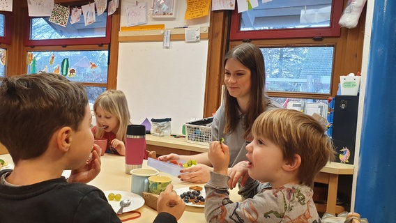 Die Auszubildende Lea Raddatz sitzt mit Kindern der Kita zusammen am Tisch und lächelt. © Johannes Tran Foto: Johannes Tran