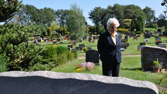 Irmgard Augustin steht vor einem Grabstein. © NDR Foto: Christian Wolf