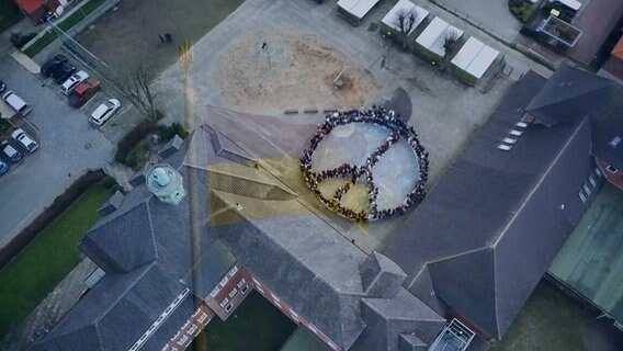 Eine Drohnenaufnahme zeigt, wie Schüler der Europaschule in Marne ein großes Peacezeichen auf ihrem Schulhof bilden. © Europaschule Marne 