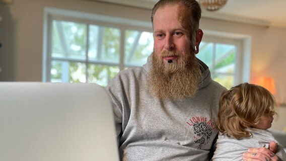 Ein bärtiger Mann hat sein Kind auf dem Arm und sitzt am Laptop. © NDR Foto: Jonas Hanst