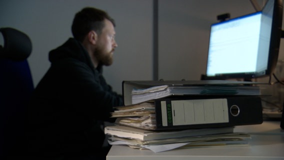 Ein Mann sitzt vor einem Computer, im Vordergrund liegt ein Aktenstapel © NDR 